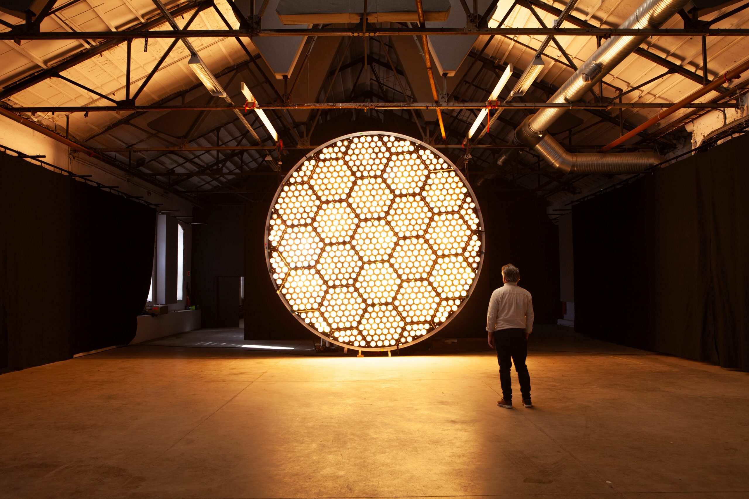 "Two people standing in front of a bright circular lighting installation, with incandescent bulbs simulating a dynamic sun effect."