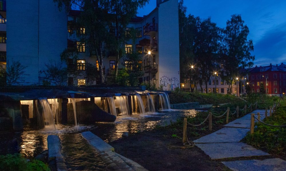 Night view of Klosterenga Park in Oslo, featuring illuminated water cascades flowing through stone structures. The park's serene lighting highlights the natural elements, with the soft glow from beneath the waterfalls complementing the urban surroundings and creating a tranquil ambiance. Trees and pathways are dimly lit in the background, blending art, nature, and lighting design seamlessly.