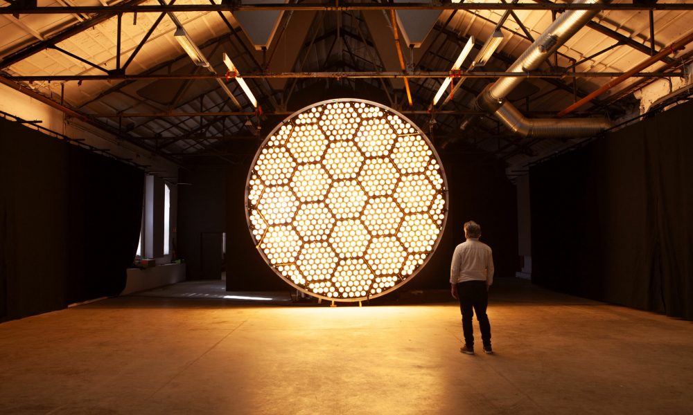 "Two people standing in front of a bright circular lighting installation, with incandescent bulbs simulating a dynamic sun effect."